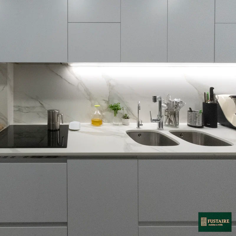 Zona de cocción y fregadero con encimera porcelánica estilo mármol e iluminación LED bajo mueble.
